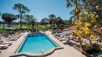 A swimming pool with lounge chairs and umbrellas, surrounded by palm trees and a golf course in the background.