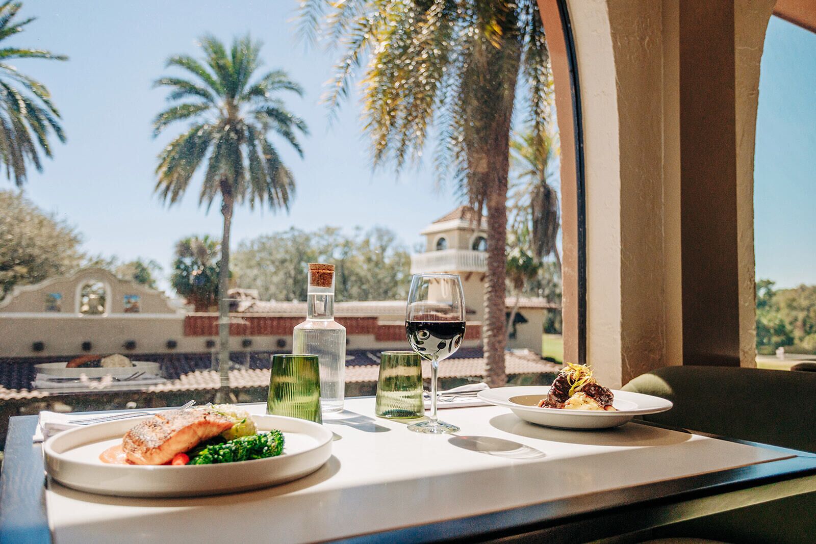 Una mesa de comedor con dos platos de comida, una botella y copas de vino, frente a una vista de palmeras y un edificio.