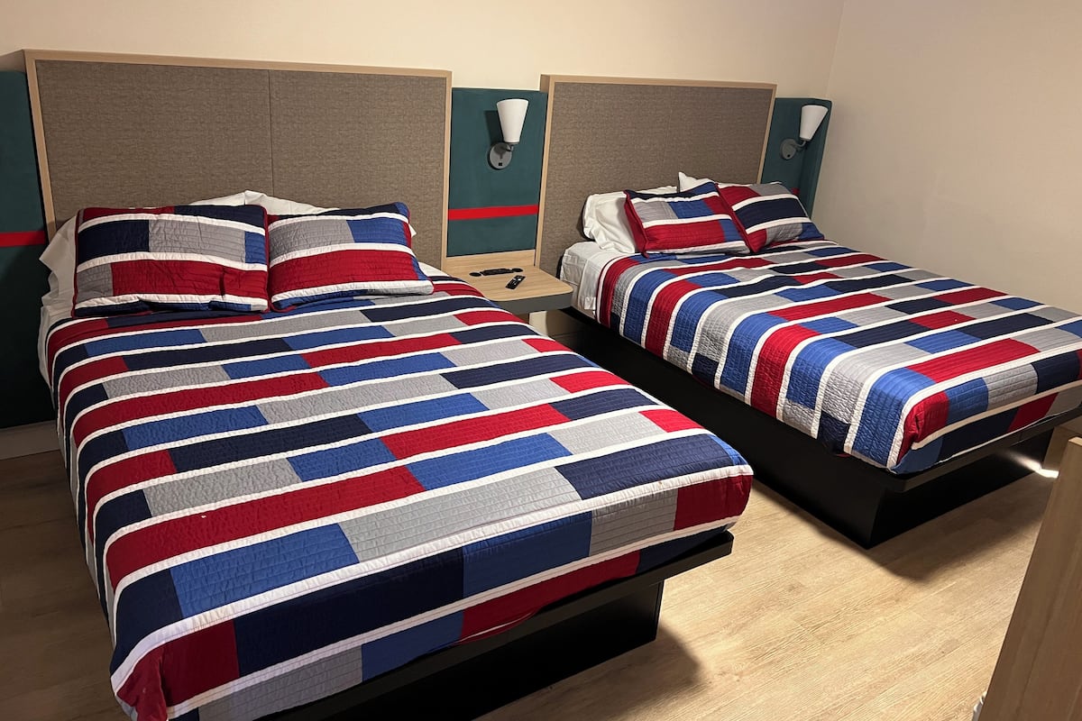 A hotel room with two beds, each with a striped bedspread and matching pillows. The room has a wooden floor and a nightstand with a lamp on each side of the beds.