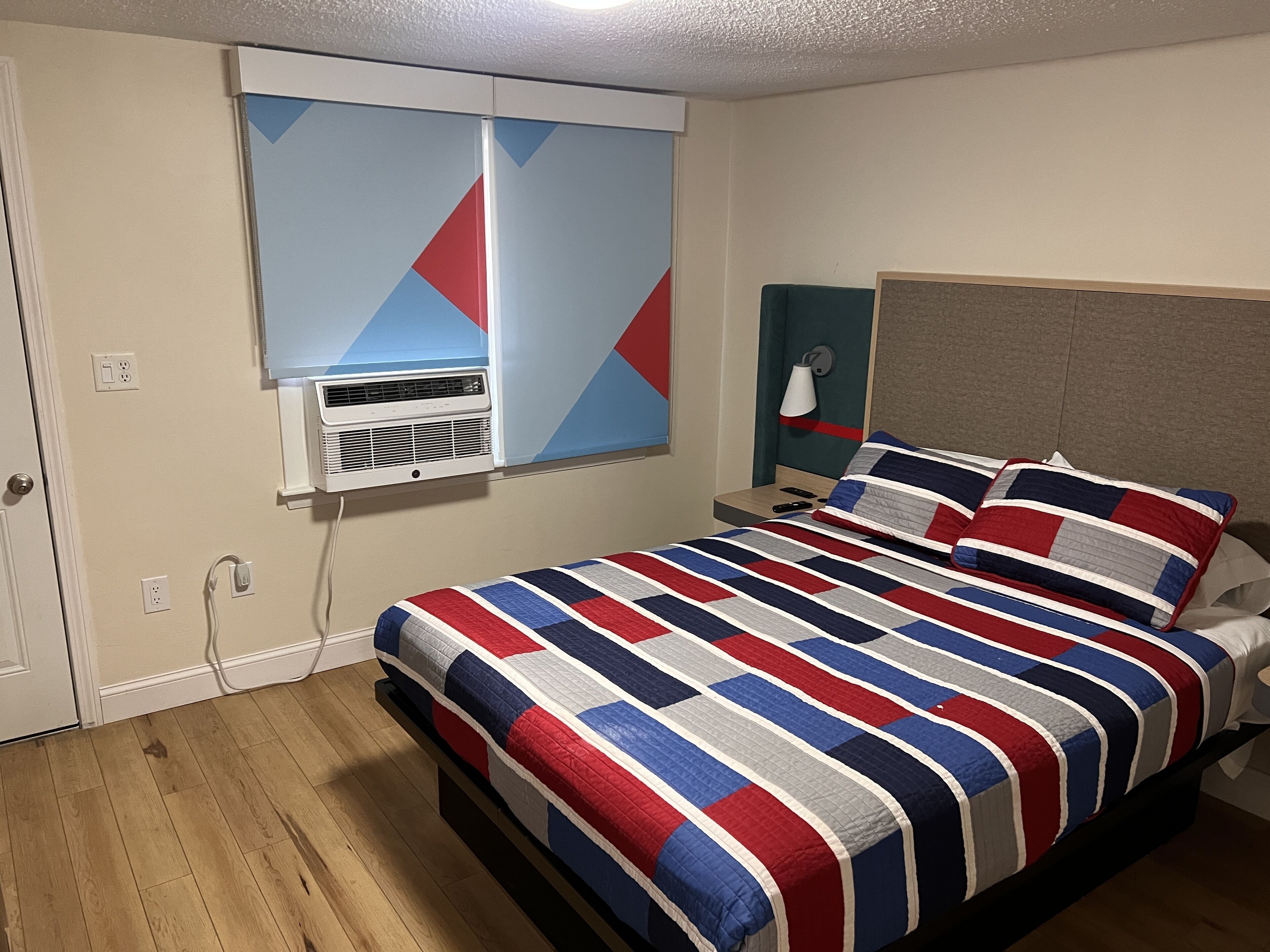 Ein Schlafzimmer mit einem Bett, einem Fenster mit Jalousien, einer Klimaanlage, einem Nachttisch mit Lampe und Holzboden.