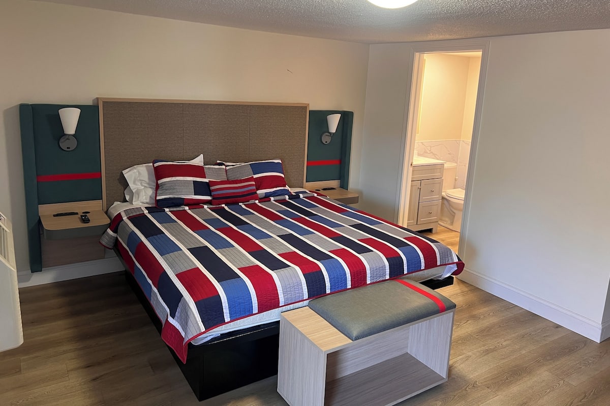 A modern bedroom with a large bed, bedside tables, a bench, and a bathroom visible through an open door.