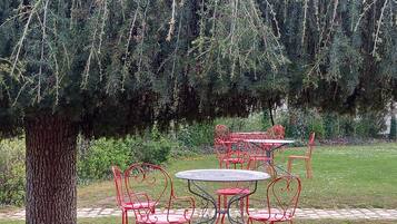 Een tuin met een grote boom, rode metalen stoelen en tafels.
