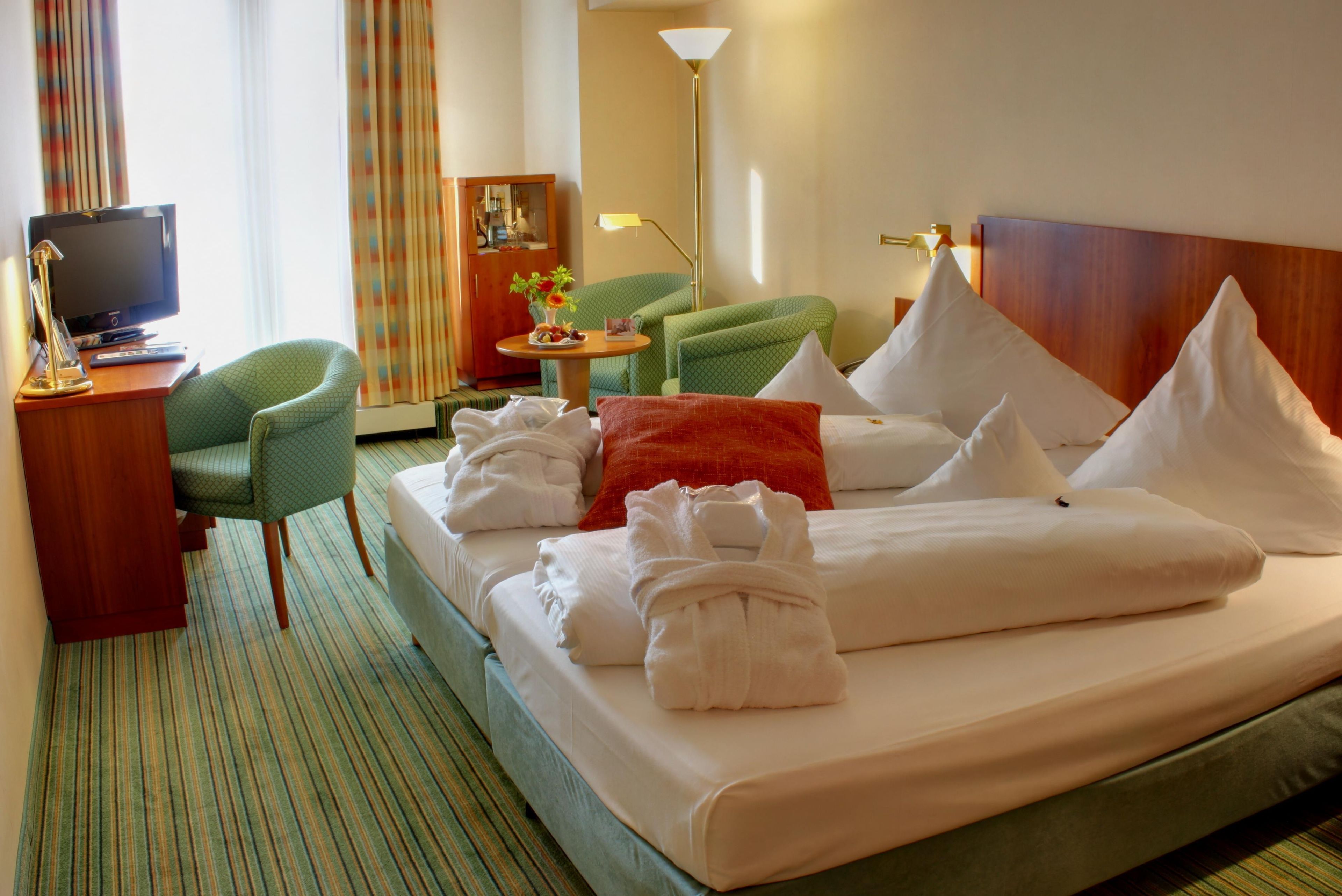 A hotel room with a bed, a desk with a television, a chair, a lamp, and a small table with a vase of flowers.