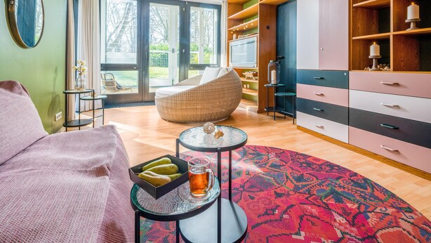 Een moderne woonkamer met een bank, een ronde salontafel met een glazen mok, een rieten stoel en een groot raam met uitzicht op bomen.