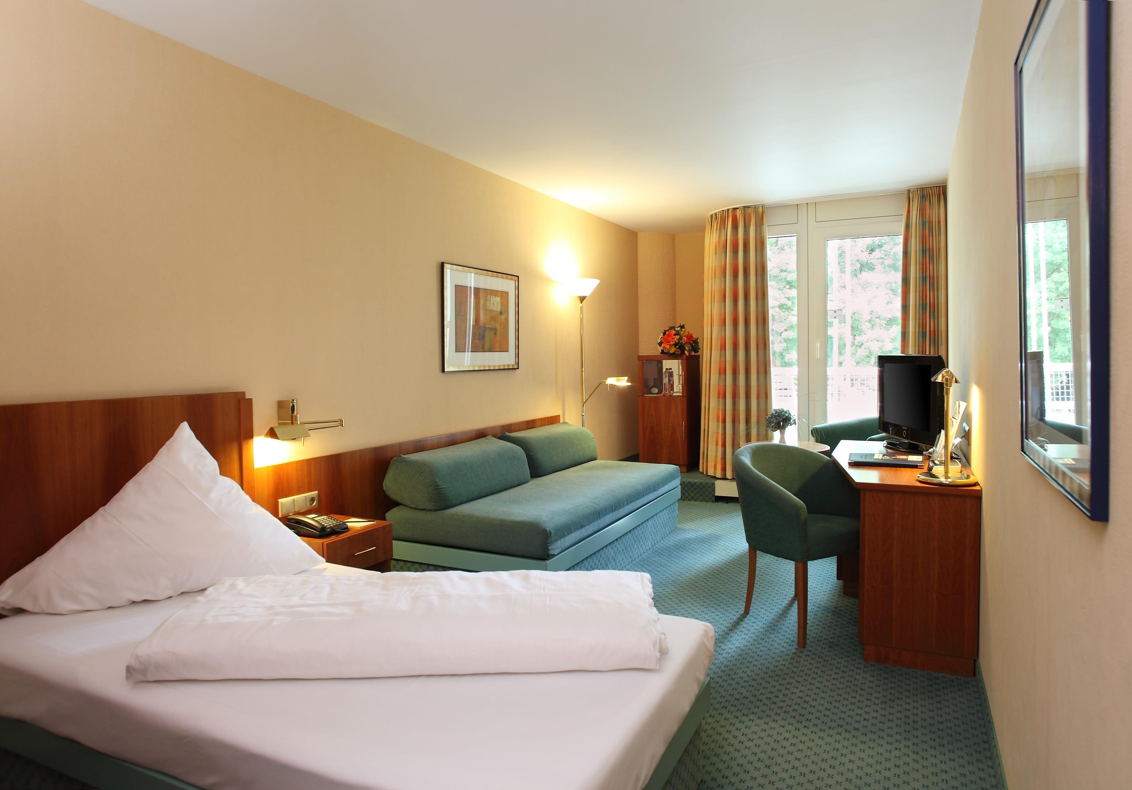 Habitación de hotel con cama, sofá, escritorio con ordenador y ventana con cortinas.
