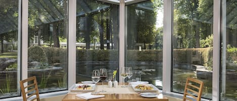 Um espaço de refeições no canto, com uma mesa posta para dois, duas cadeiras e vista para um jardim através de grandes janelas.