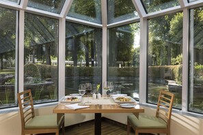 A corner dining area with a table set for two, two chairs, and a view of a garden through large windows. - Best Western Premier Parkhotel Bad Mergentheim (Bad Mergentheim)