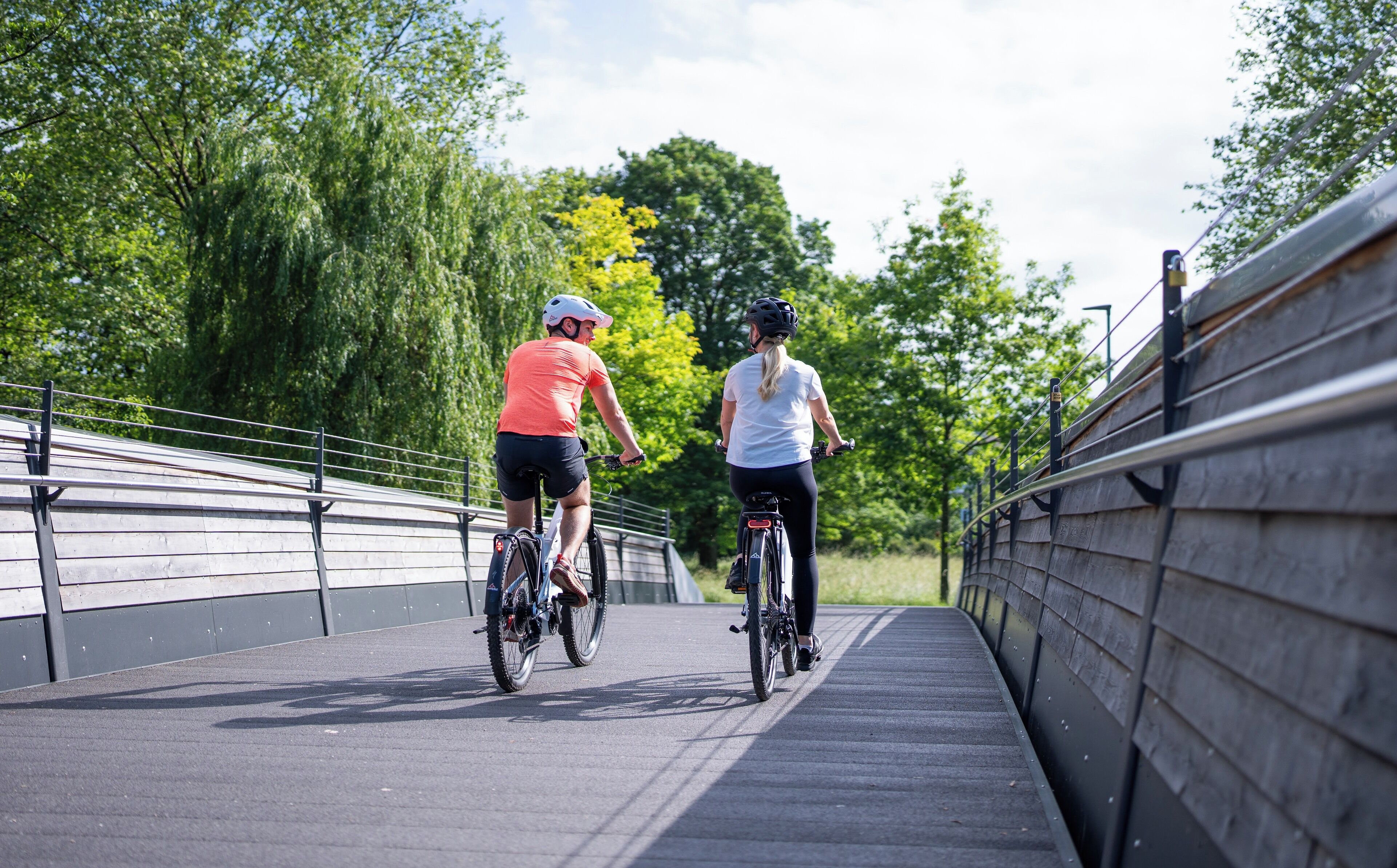 Two cyclists on a paved path with metal railings on both sides, surrounded by greenery.