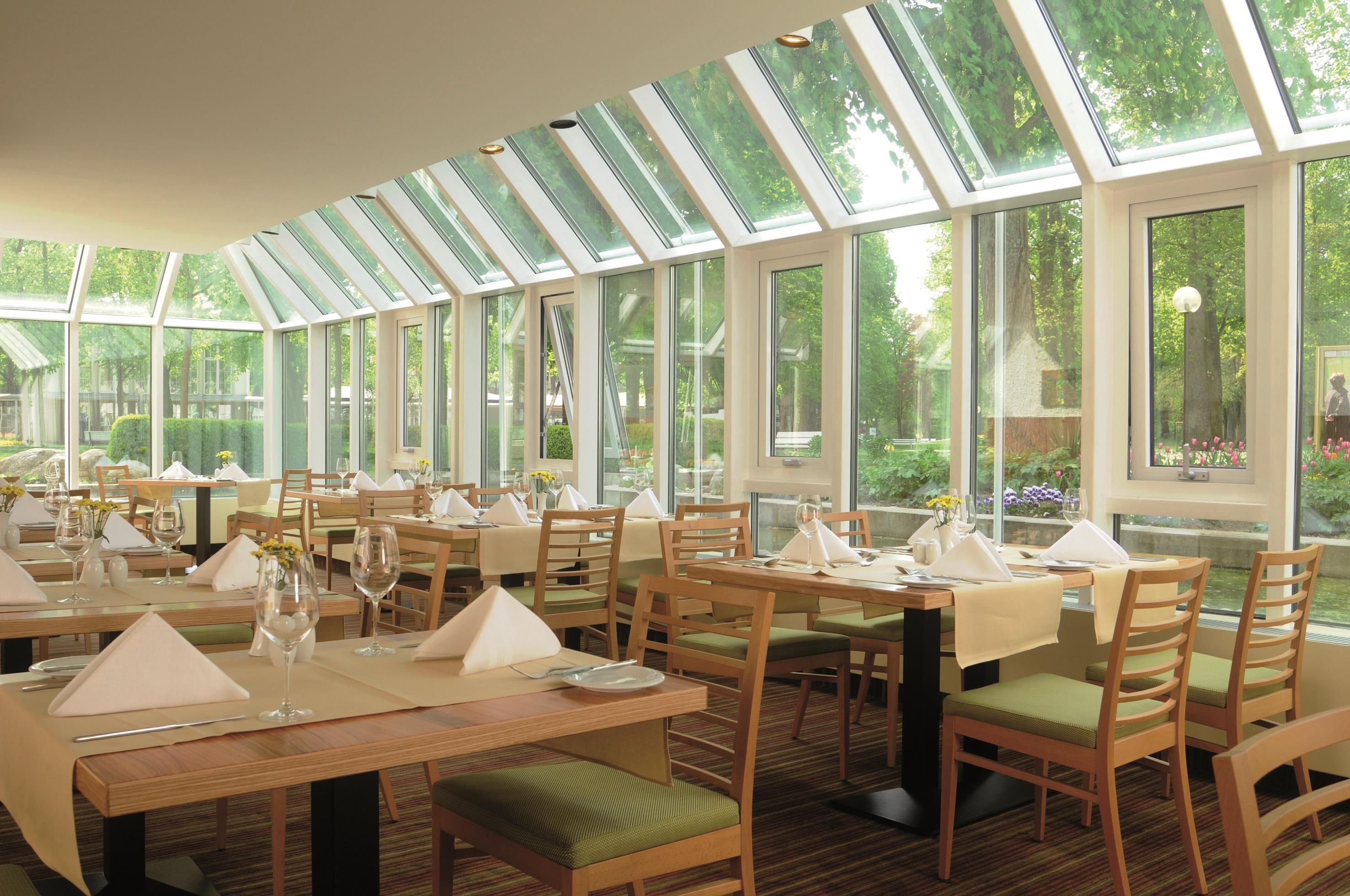 Un restaurante con varias mesas, cada una dispuesta con servilletas blancas, vasos y cubiertos. El salón cuenta con grandes ventanales que dejan entrar luz natural y ofrecen vistas a la vegetación exterior.