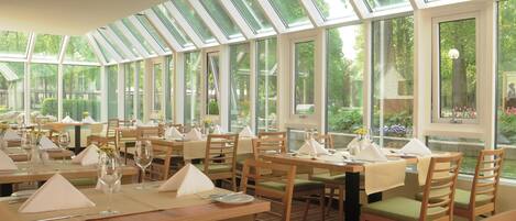 Un restaurante con varias mesas, cada una dispuesta con servilletas blancas, vasos y cubiertos. El salón cuenta con grandes ventanales que dejan entrar luz natural y ofrecen vistas a la vegetación exterior.