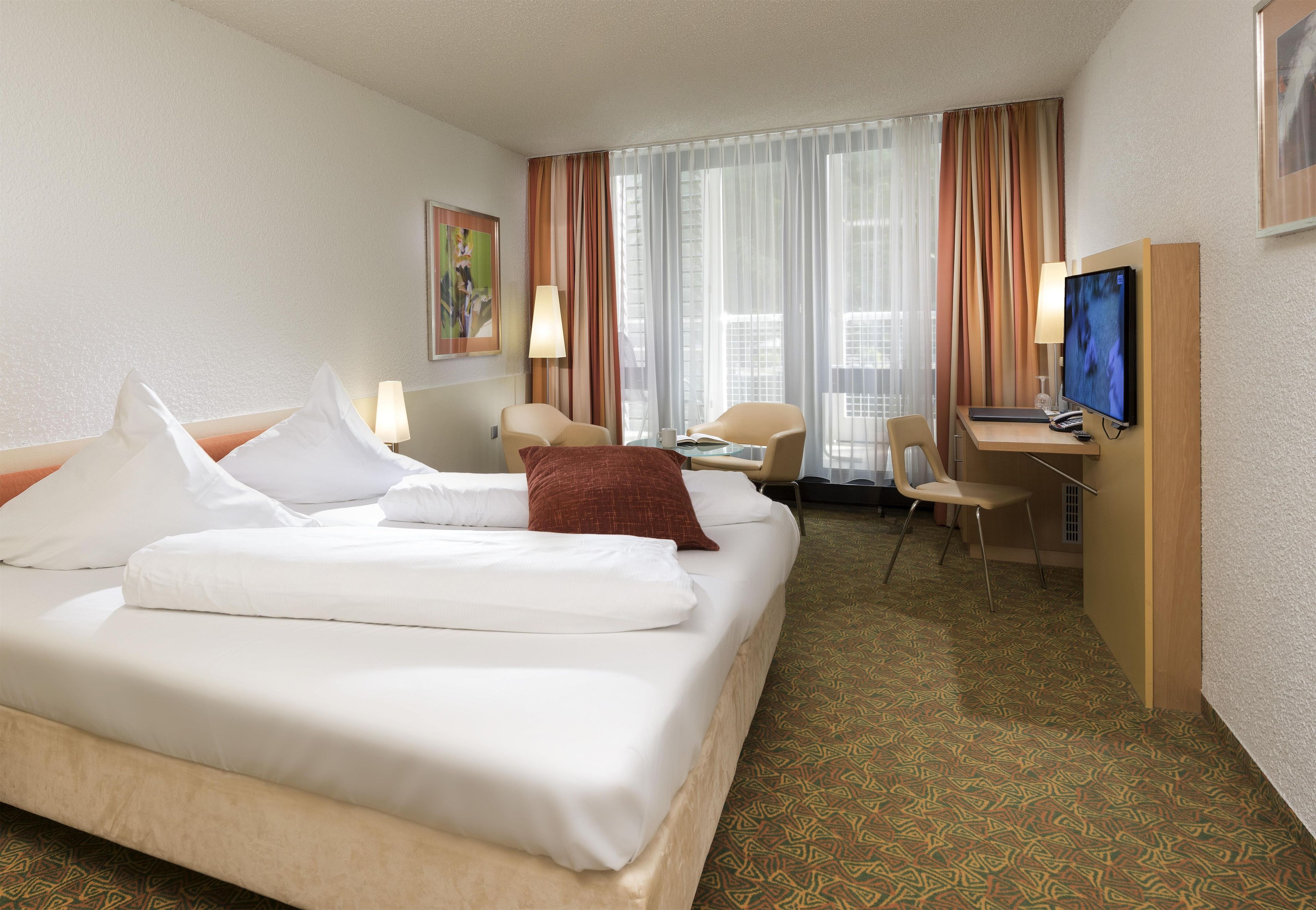 Ein Hotelzimmer mit Bett, Schreibtisch, Stuhl, Fernseher und einem Fenster mit Vorhängen.