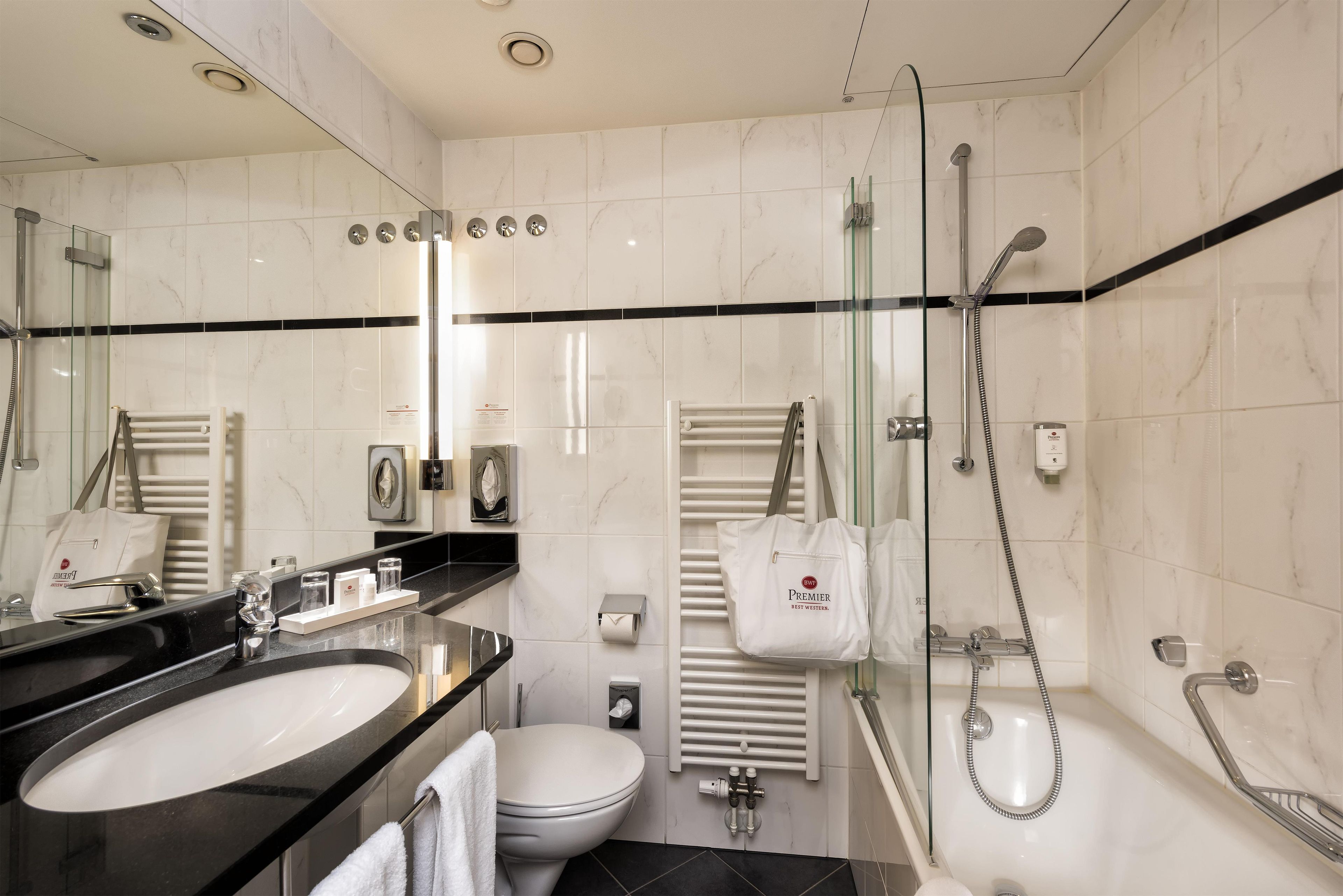 Un baño moderno con lavamanos sobre encimera negra, inodoro blanco, ducha con mampara de cristal y toallero con una bolsa blanca.