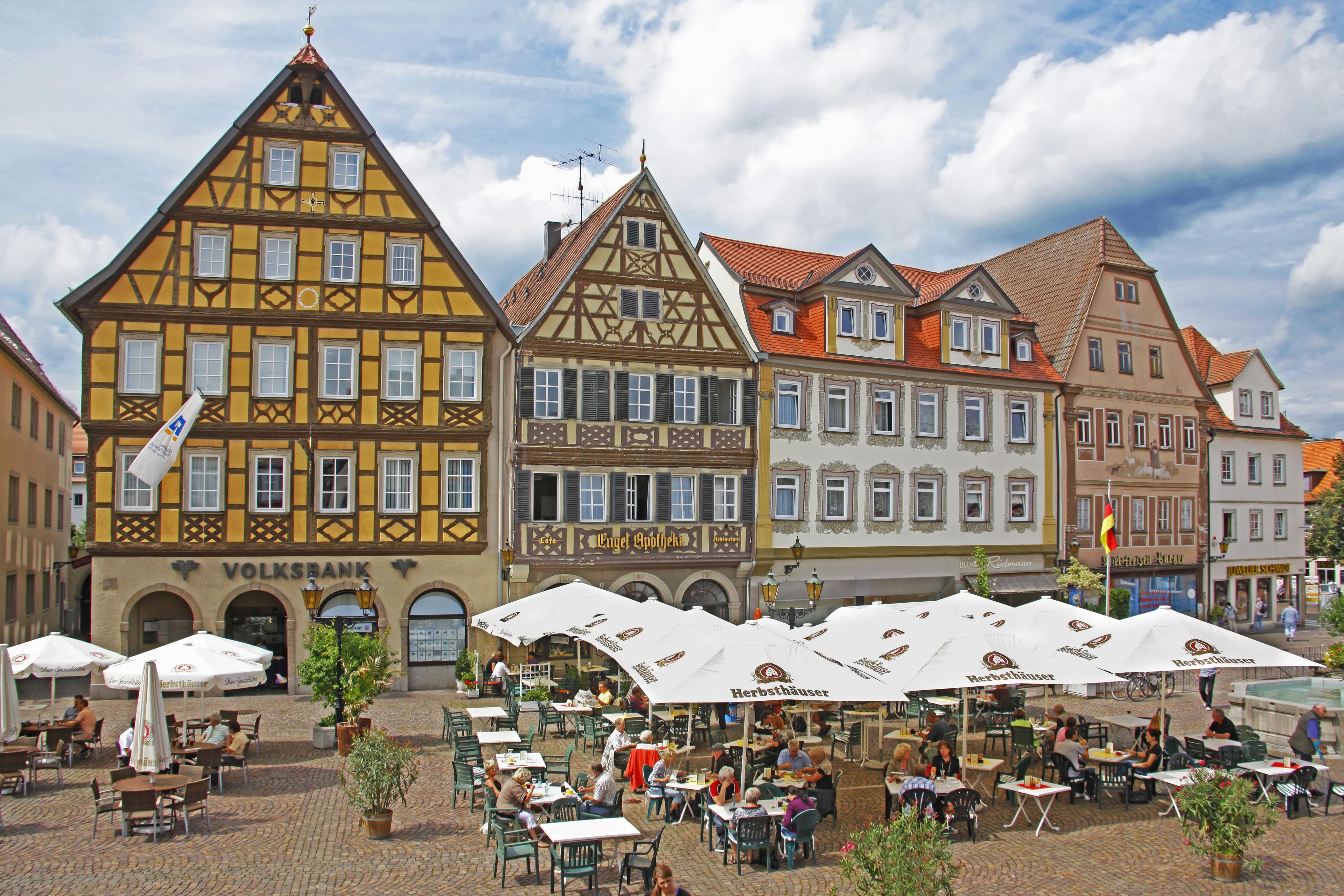 Une place typique d’une ville européenne, avec des bâtiments à colombages, des terrasses et une succursale de la banque Volksbank.