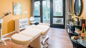 Een behandelruimte met een massagetafel, een plank met flesjes, een spiegel en een raam met uitzicht op bomen.
