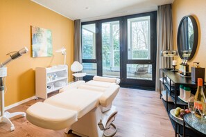 A treatment room with a massage table, a shelf with bottles, a mirror, and a window with a view of trees.