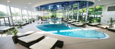 An indoor swimming pool with lounge chairs and potted plants, surrounded by large windows.