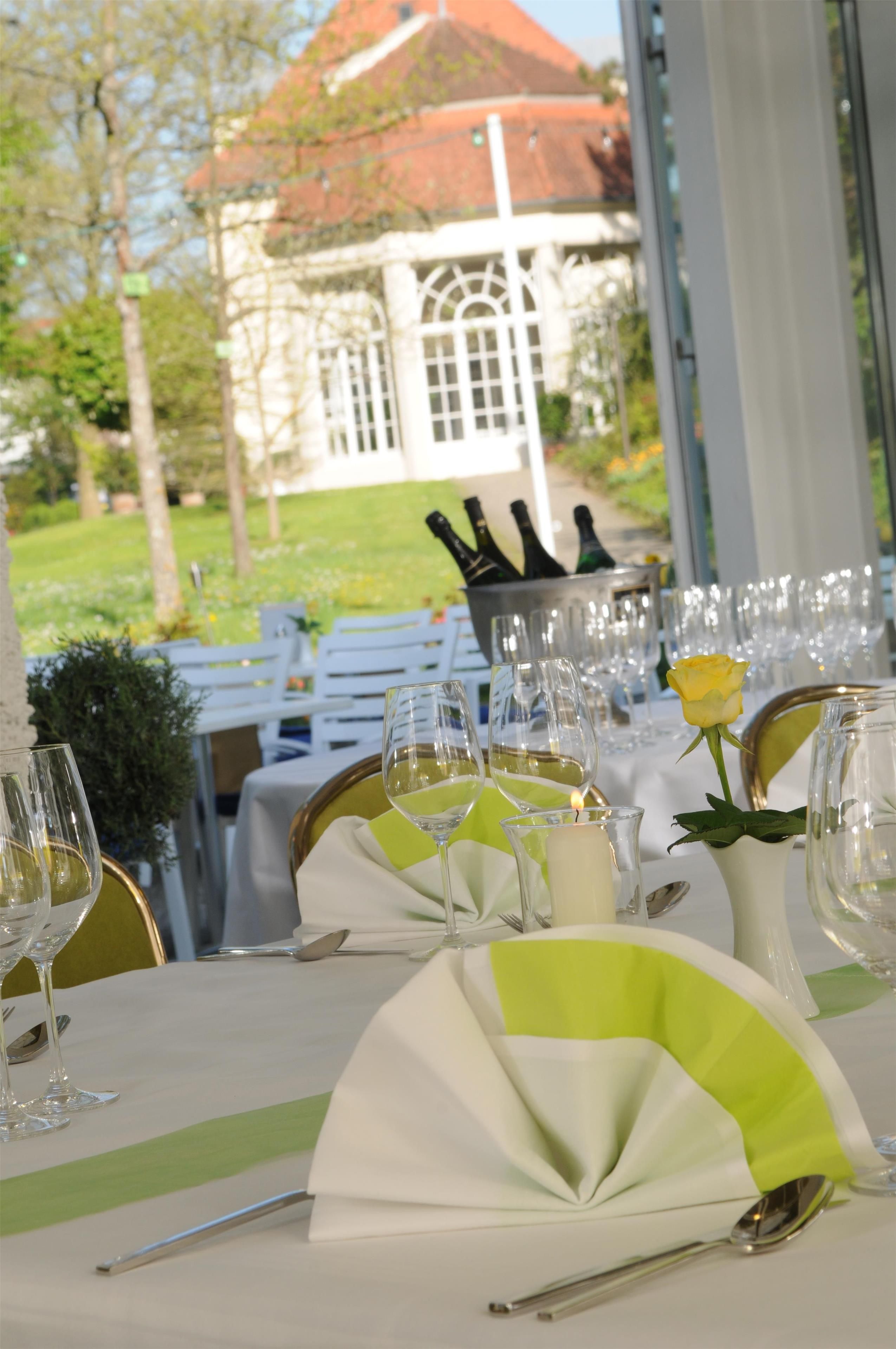 An outdoor dining area with tables set for a meal, a view of a building with a red roof, and a rose in a vase.