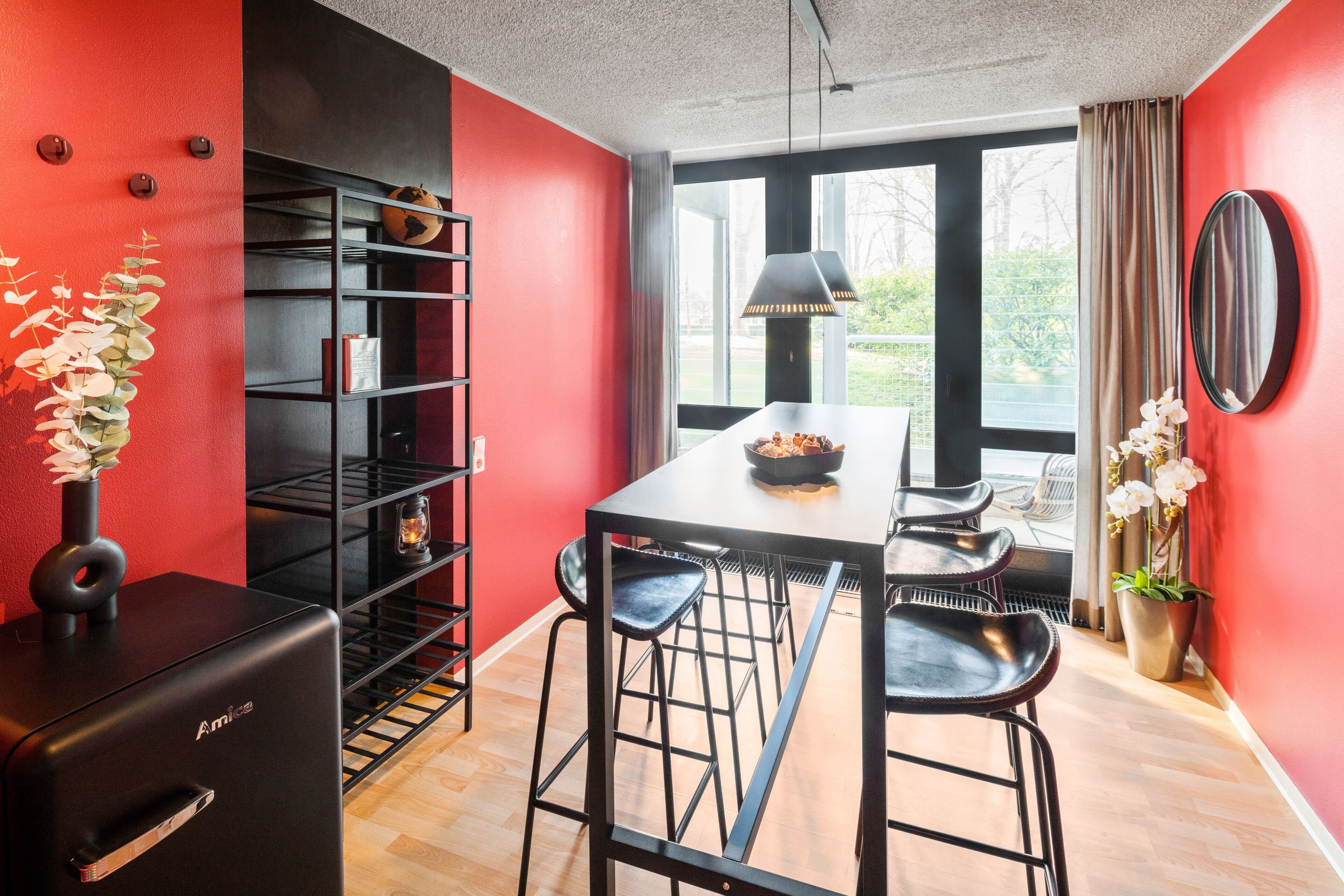 A modern kitchen with a black refrigerator, a dining table with bar stools, and a large window with a view of greenery.