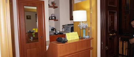 Un mostrador de recepción con un teléfono, una lámpara y un jarrón de flores. Hay un espejo sobre el mostrador y una puerta a la izquierda.