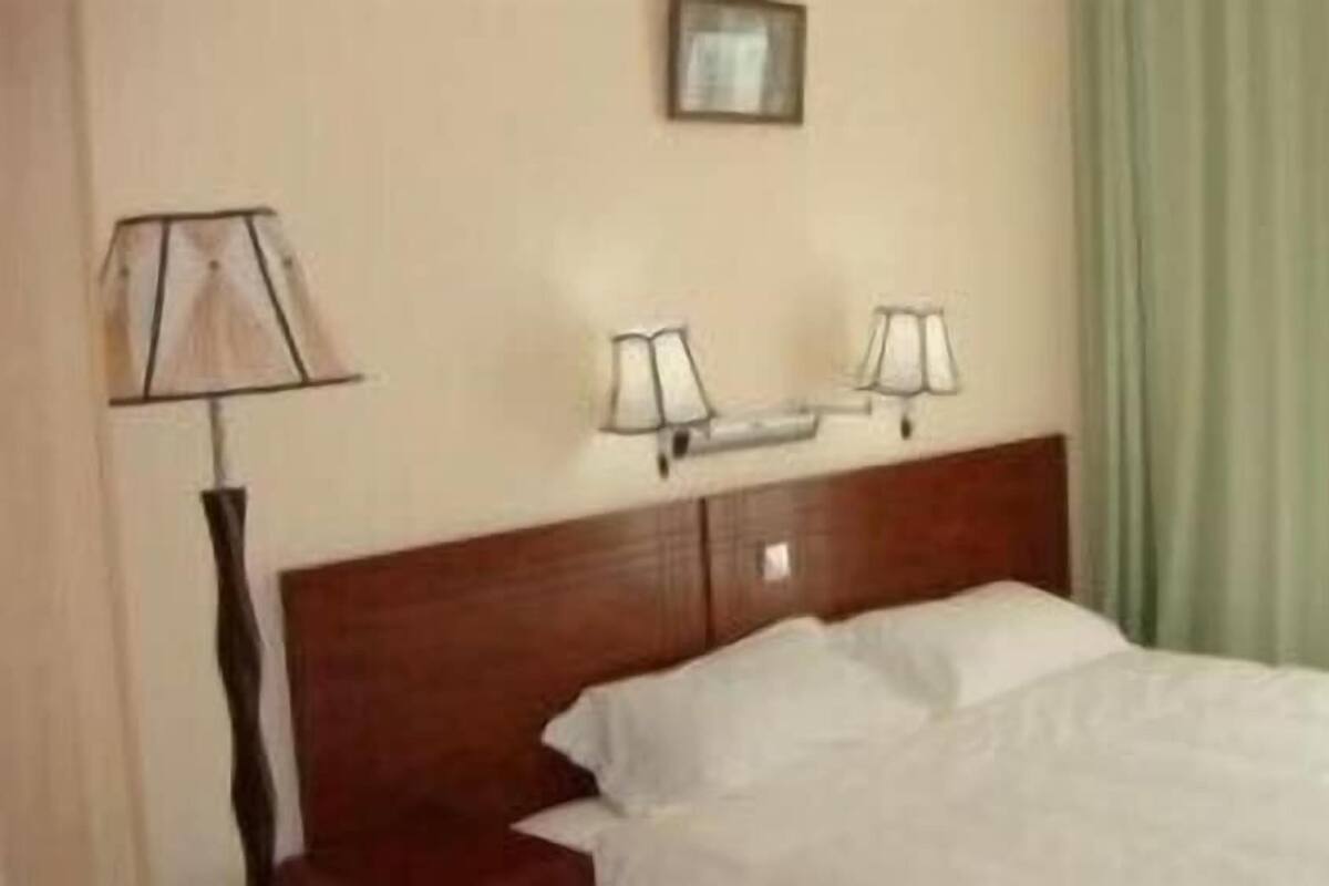 Hotellihuone, jossa on puinen sängynpääty, kaksi yöpöytävalaisinta, valkoisilla lakanoiden peitetty sänky ja vihreä verho.