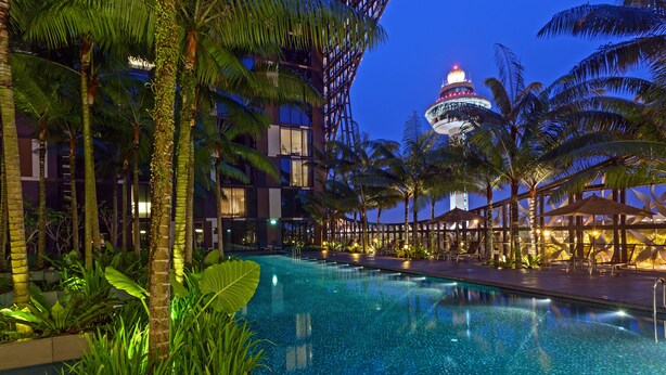 Zona de piscina con palmeras, un edificio con estructura en forma de cúpula y un paseo bien iluminado.