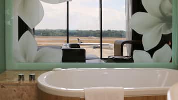 Un baño con una bañera grande, un espejo que refleja un avión en la pista y vistas al aeropuerto.