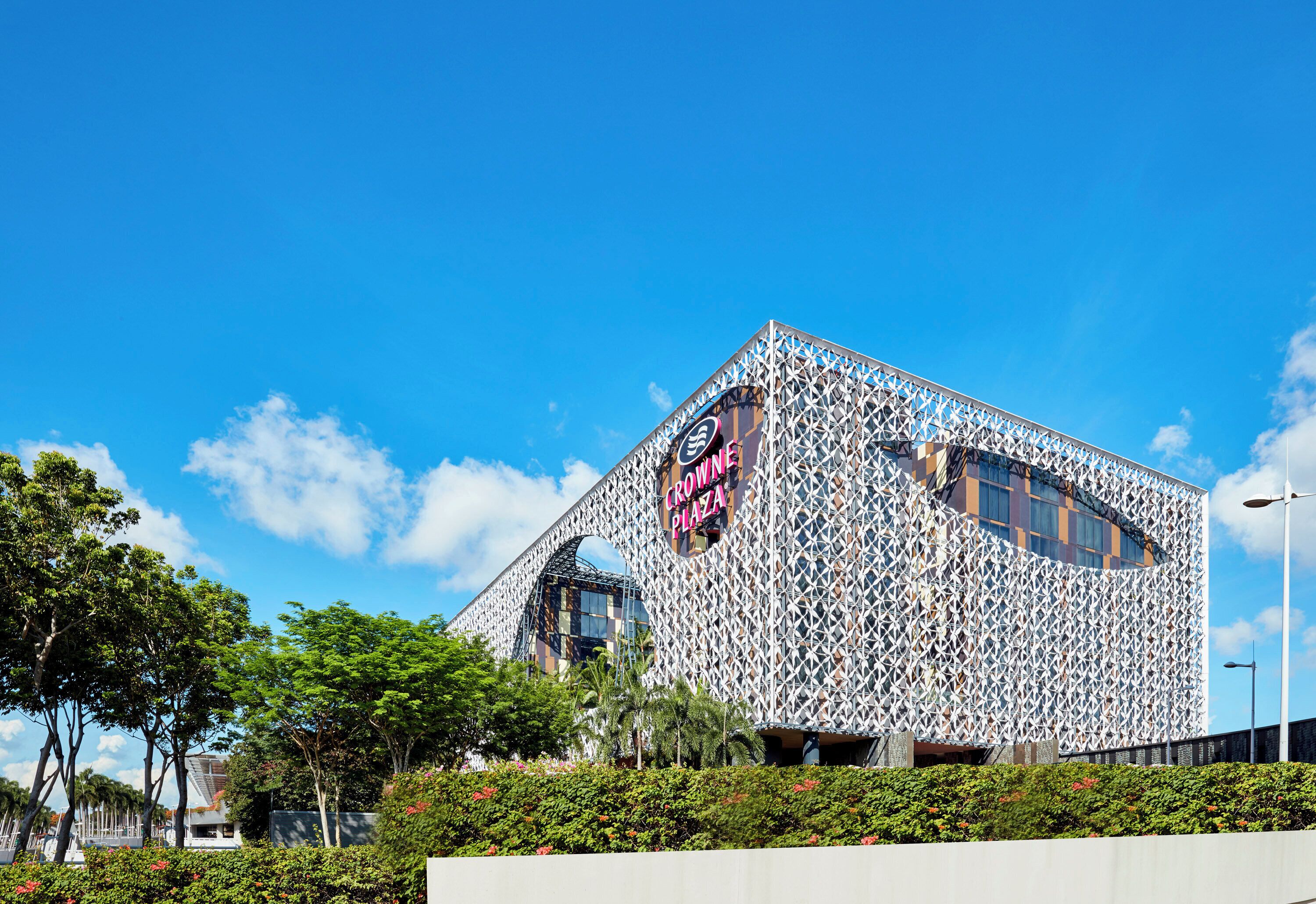 A modern building with a distinctive geometric facade, a prominent sign reading 'CROWNE PLAZA,' and lush greenery in the foreground.