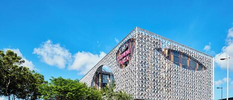 A modern building with a distinctive geometric facade, a prominent sign reading 'CROWNE PLAZA,' and lush greenery in the foreground.