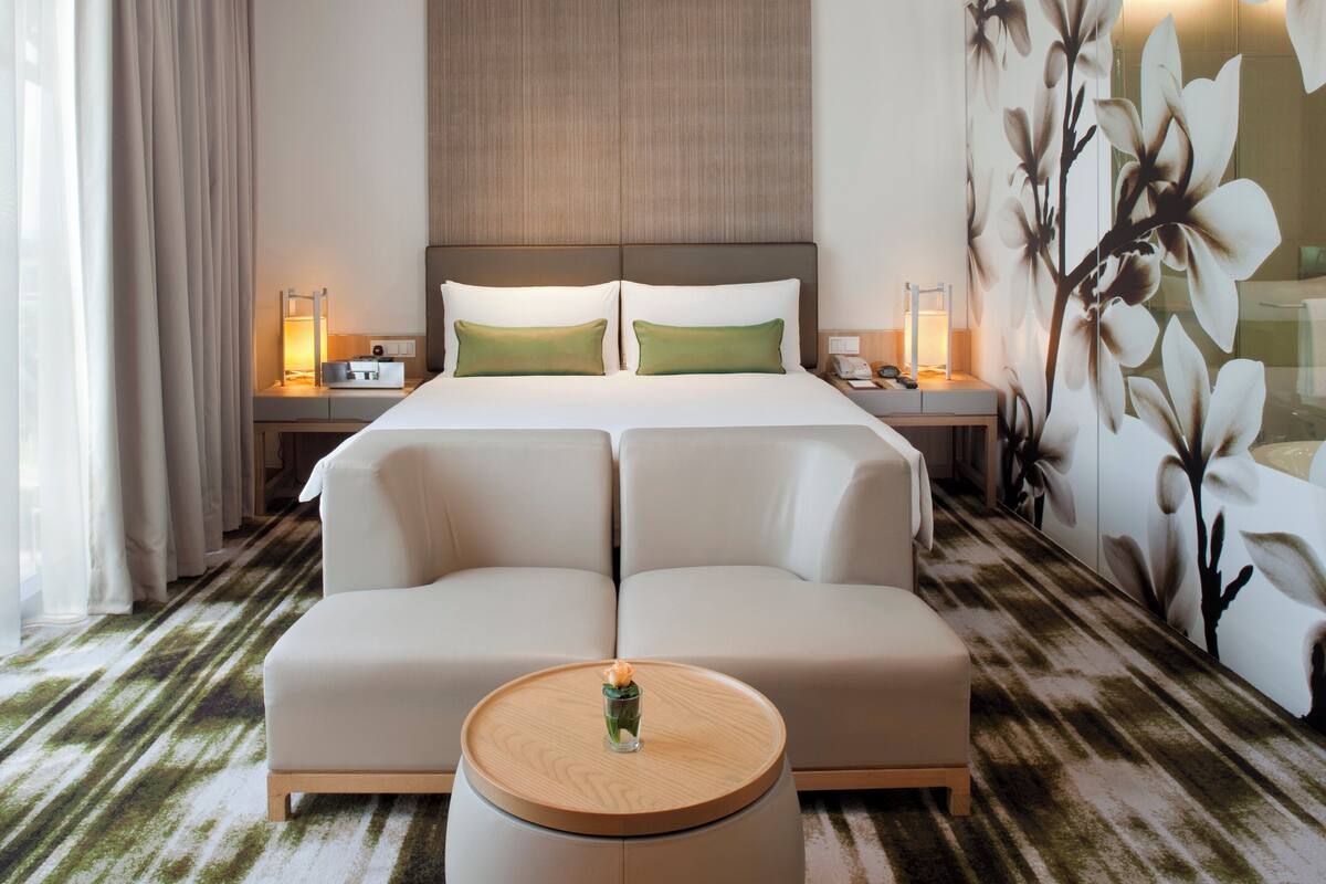 Habitación de hotel con una cama grande, dos mesitas de noche, una mesita auxiliar redonda de madera y un mural floral en la pared.