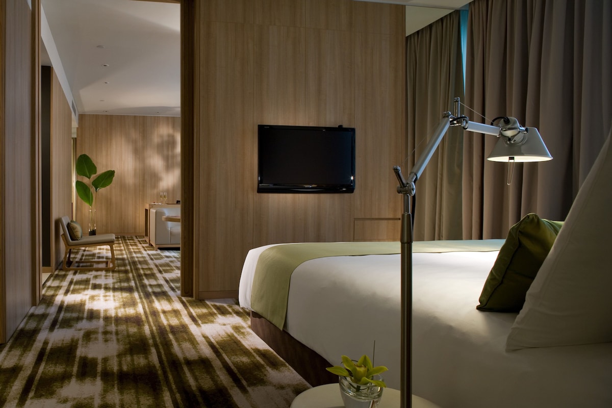 A hotel room with a bed, a bedside table with a lamp, a TV mounted on the wall, and a carpeted hallway.