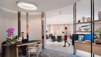A modern office space with a reception desk, two seated individuals working on laptops, a walking person, and a dining area in the background.
