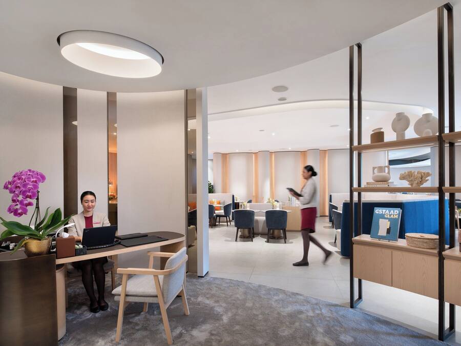 A modern office space with a reception desk, two seated individuals working on laptops, a walking person, and a dining area in the background.