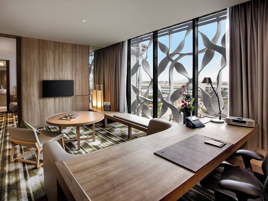 Quarto de hotel com uma janela grande, televisão de tela plana na parede, mesa redonda com cadeiras, escrivaninha com um laptop e um telefone.