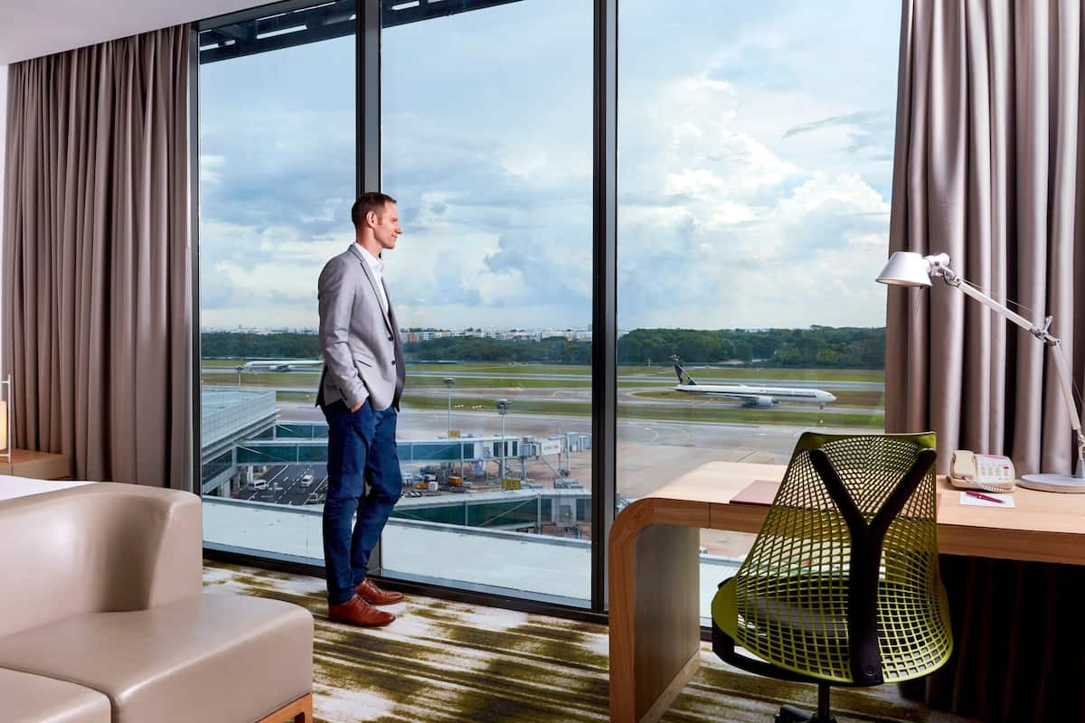 Habitación de hotel con un ventanal grande que ofrece vistas al aeropuerto, una cama, un escritorio con lámpara y una silla.