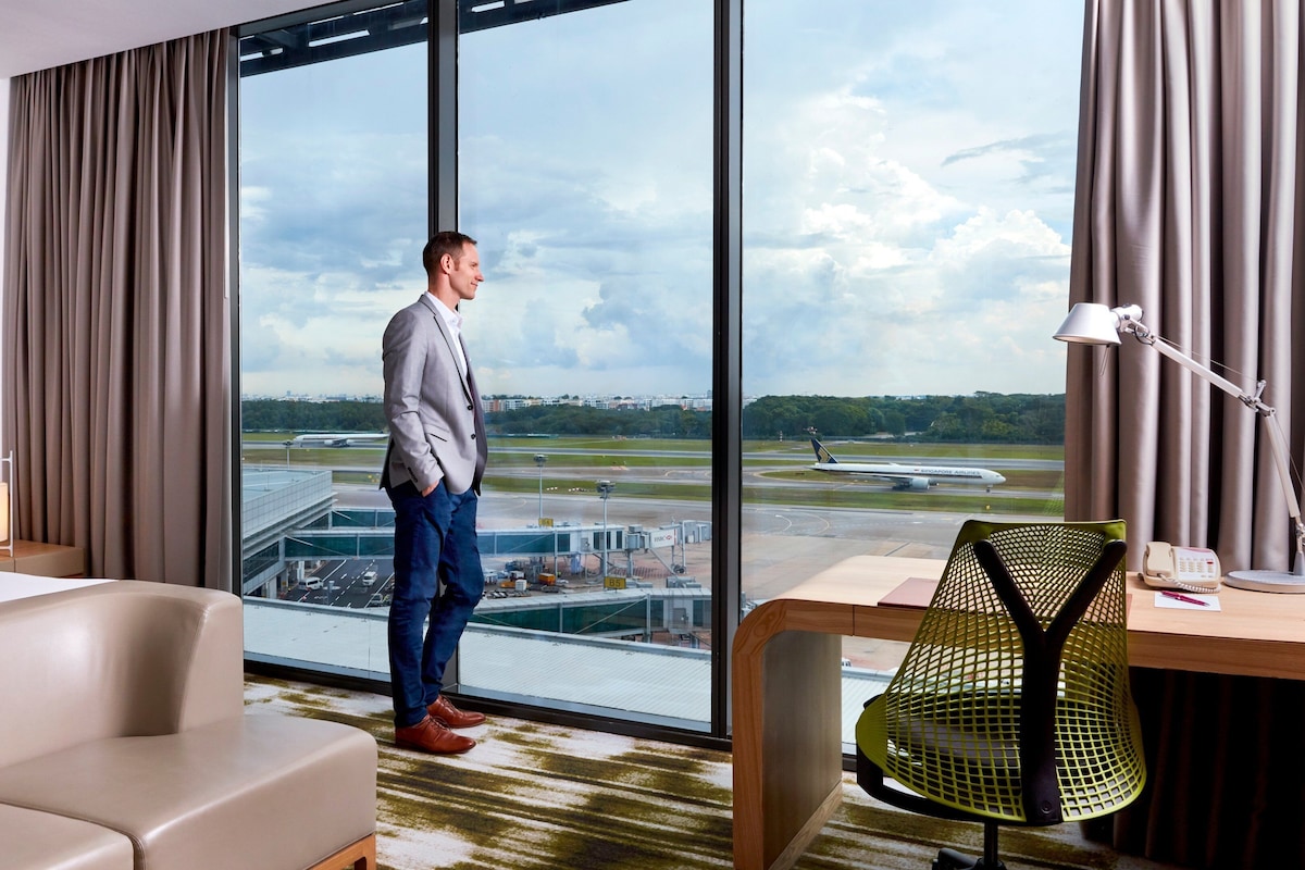 A hotel room with a large window offering a view of an airport, a bed, a desk with a lamp, and a chair.