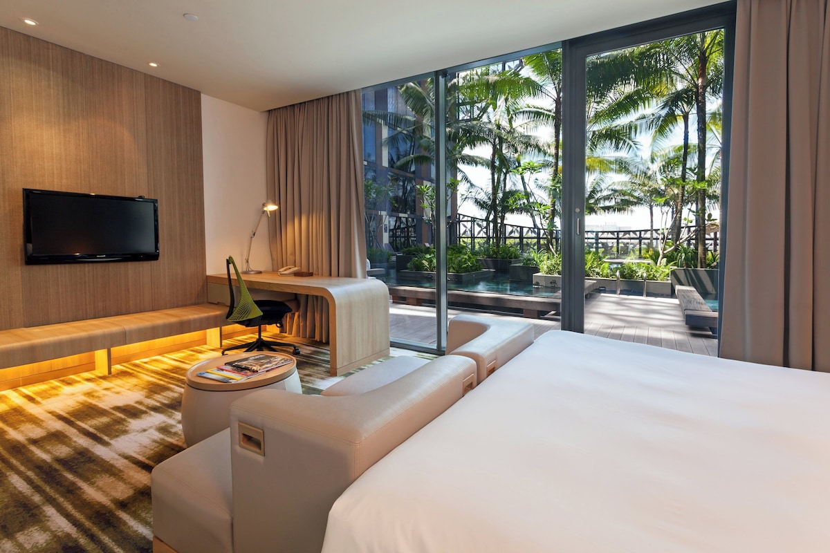 A modern hotel room with a large bed, a desk, and a TV. It has a large window with a view of palm trees and a pool.
