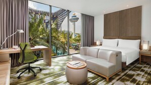 Habitación de hotel con un ventanal grande, una cama, un escritorio con silla, un taburete redondo pequeño y una mesita de noche con lámpara.