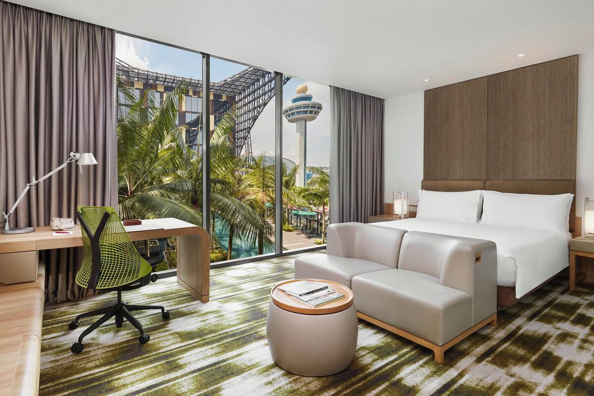 Habitación de hotel con un ventanal grande, una cama, un escritorio con silla, un taburete redondo pequeño y una mesita de noche con lámpara.