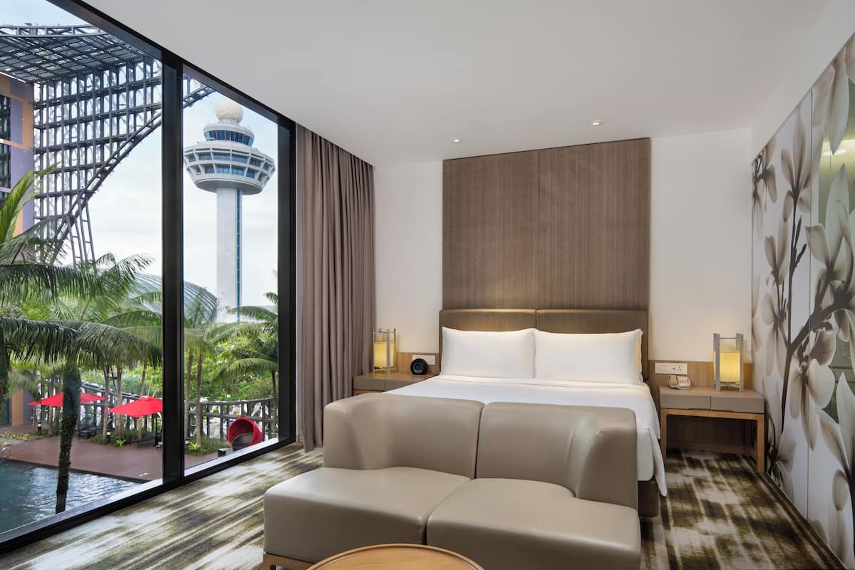 Habitación de hotel con un ventanal grande, una cama, un sofá, una mesita de noche y vistas a una torre y palmeras.