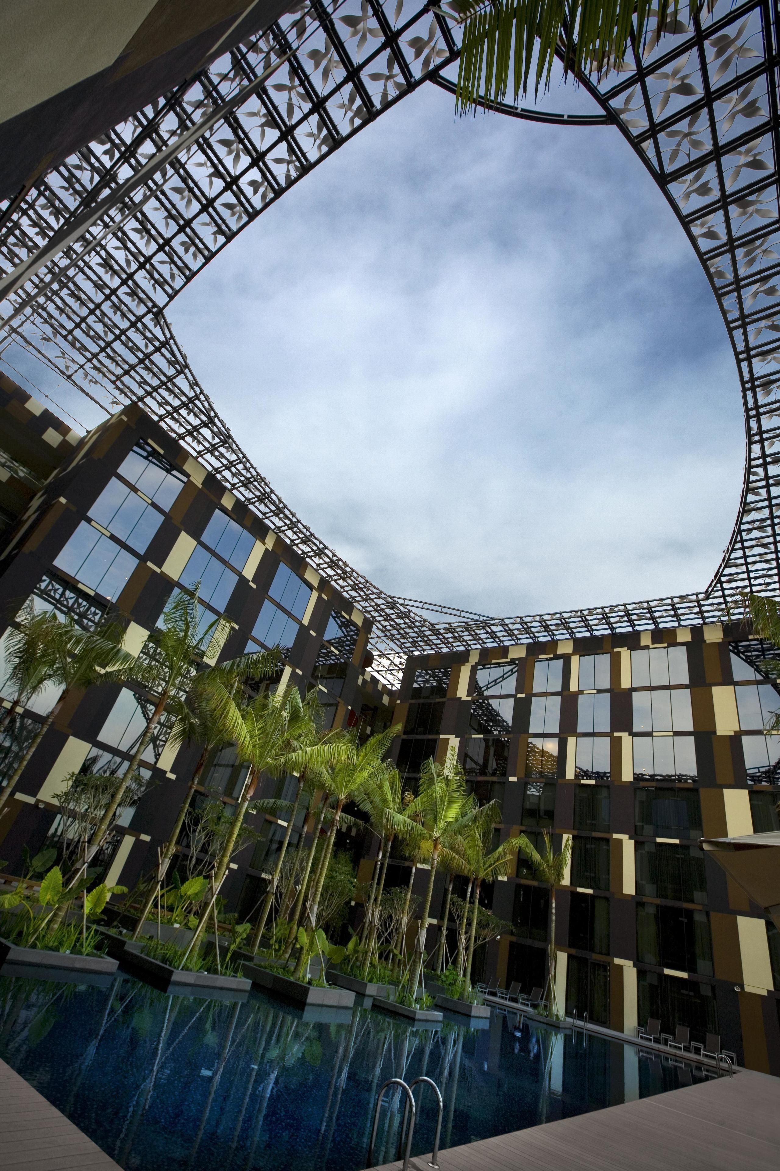 A modern building with a curved glass facade, a pool area with palm trees, and a clear sky.
