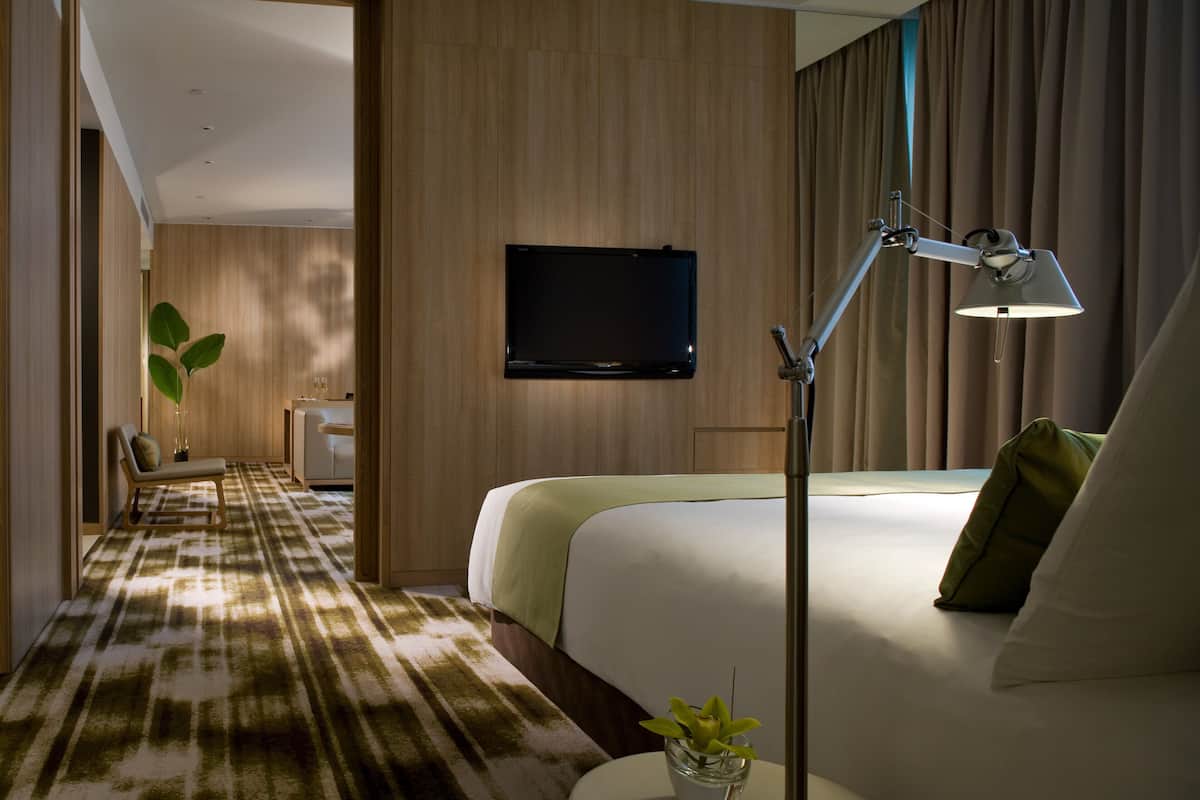 Habitación de hotel con cama, mesita de noche con lámpara, televisor montado en la pared y pasillo cubierto de alfombra.