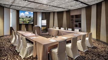 A conference room with a long table, chairs, a projector screen, and a view of a cityscape through large windows.