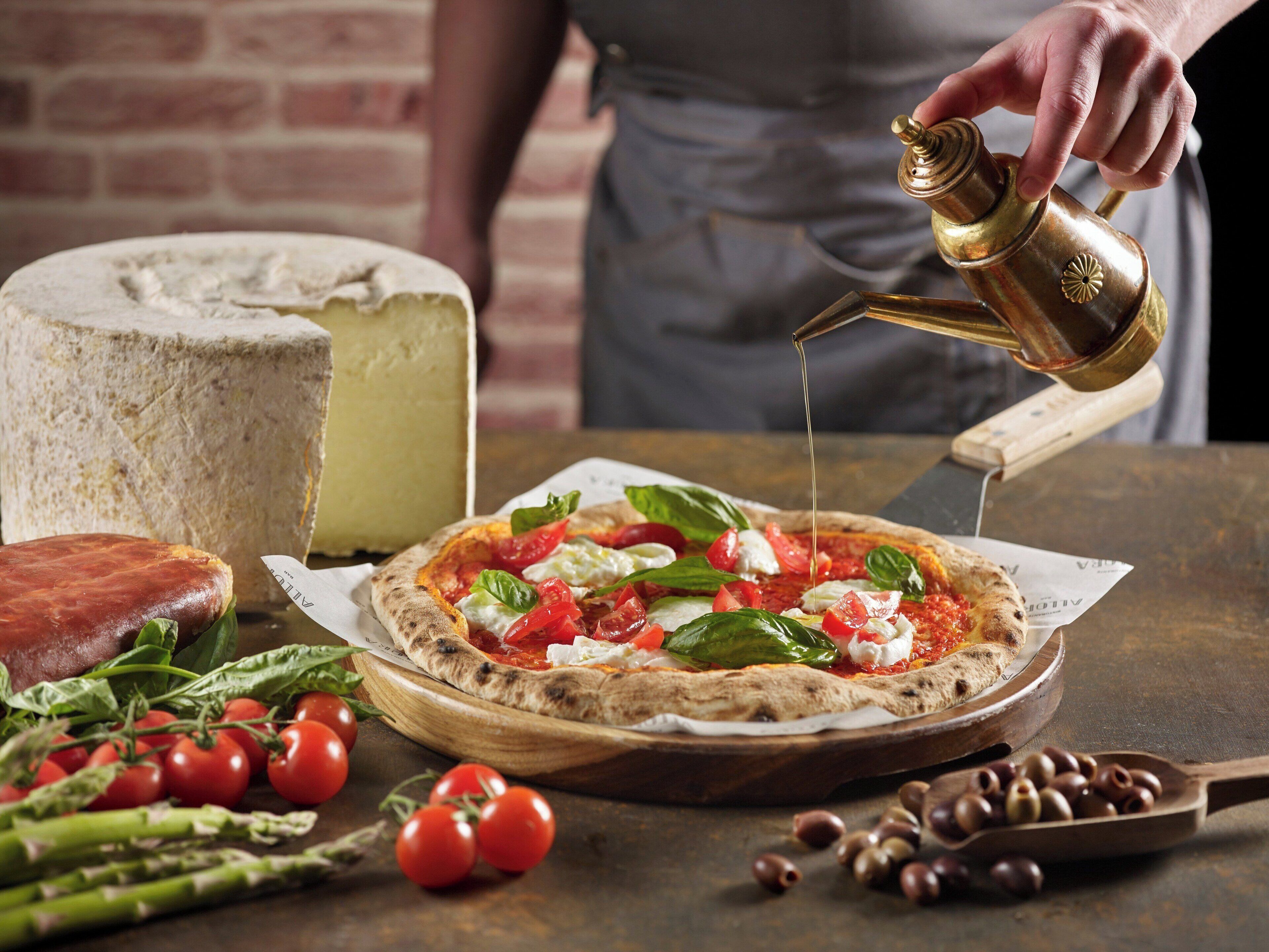 A pizza with toppings being drizzled with sauce, a wheel of cheese, and fresh vegetables on a wooden table.