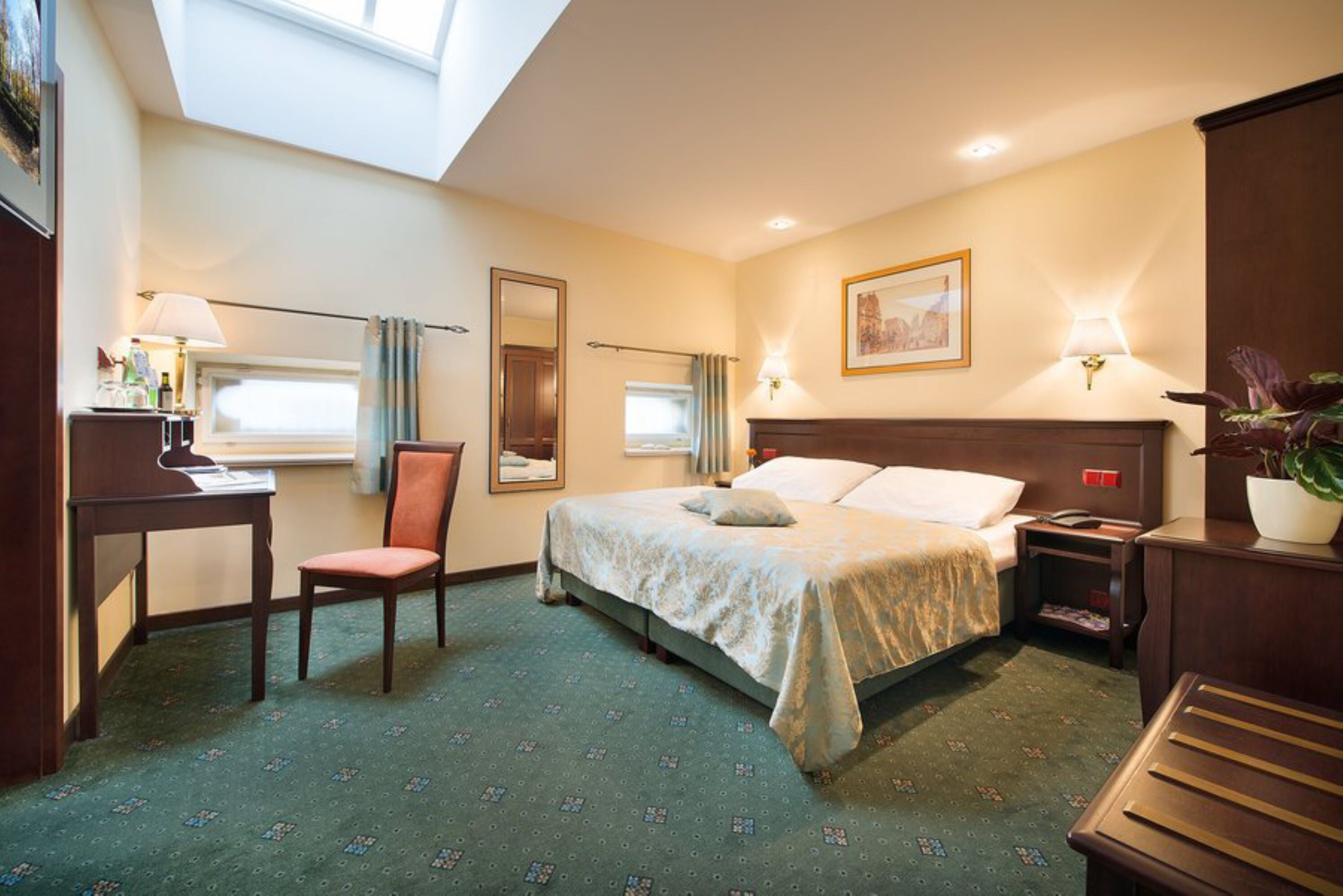 Habitación de hotel con cama, escritorio, silla, mesita de noche, lámpara, espejo y ventana.
