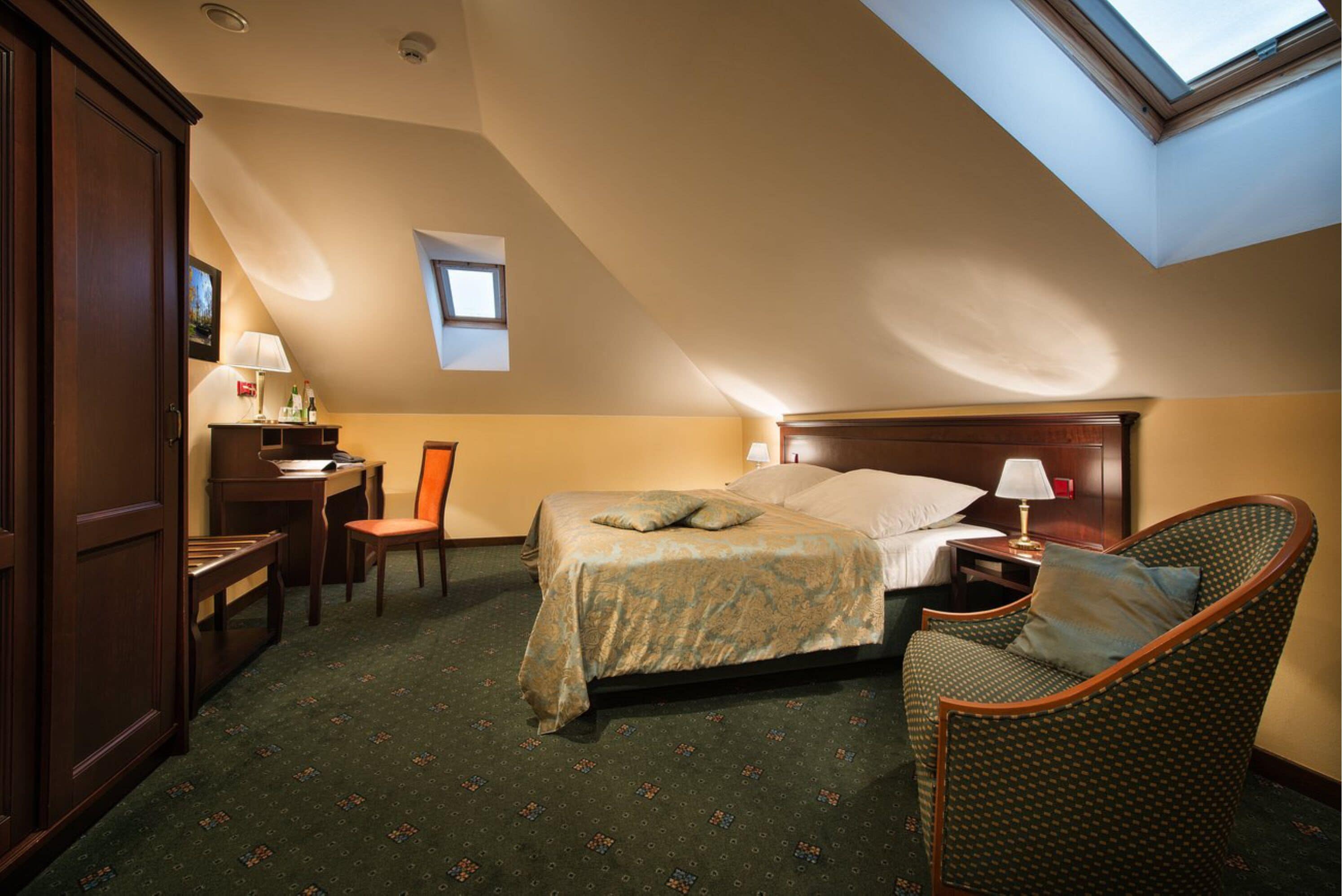 A hotel room with a bed, a chair, a desk, a lamp, and a small window.