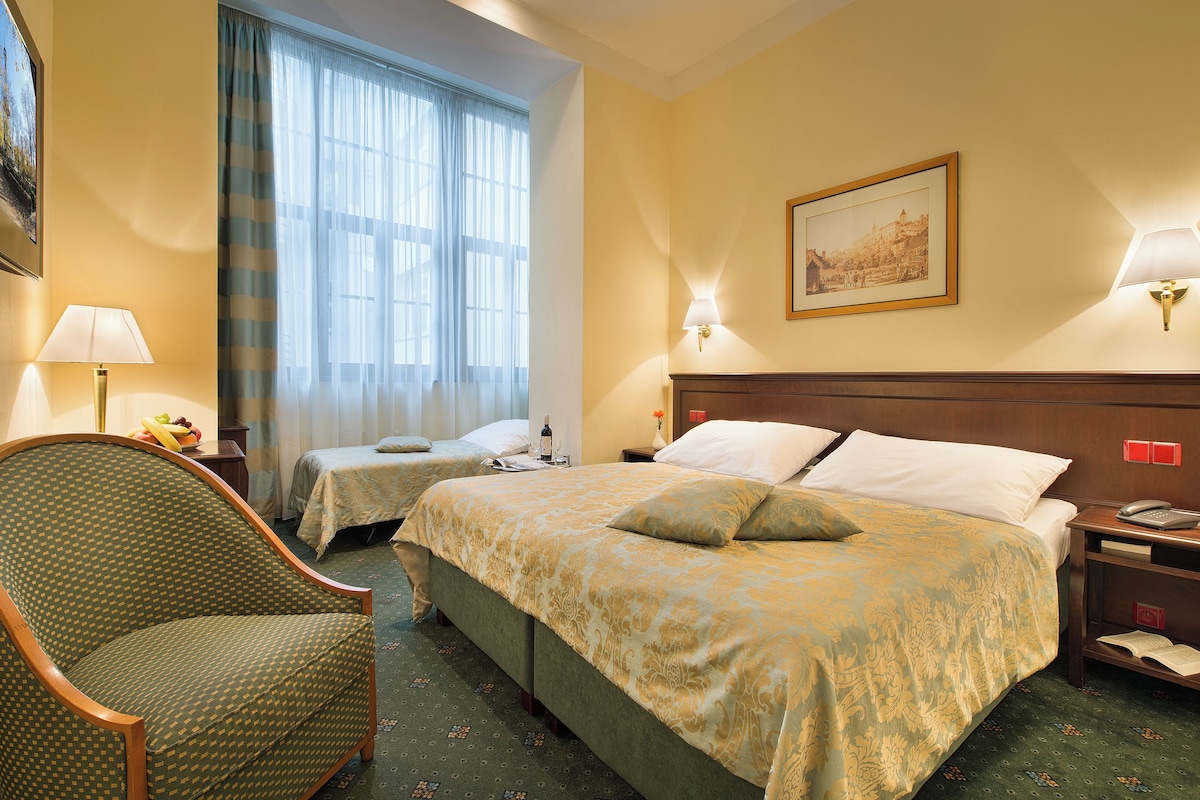 A hotel room with two beds, a chair, a lamp, a painting, and a nightstand with a phone.