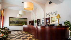 A hotel reception area with a checkered floor, a wooden reception desk, multiple clocks, and a large landscape painting. - EA Hotel Downtown (Prague)