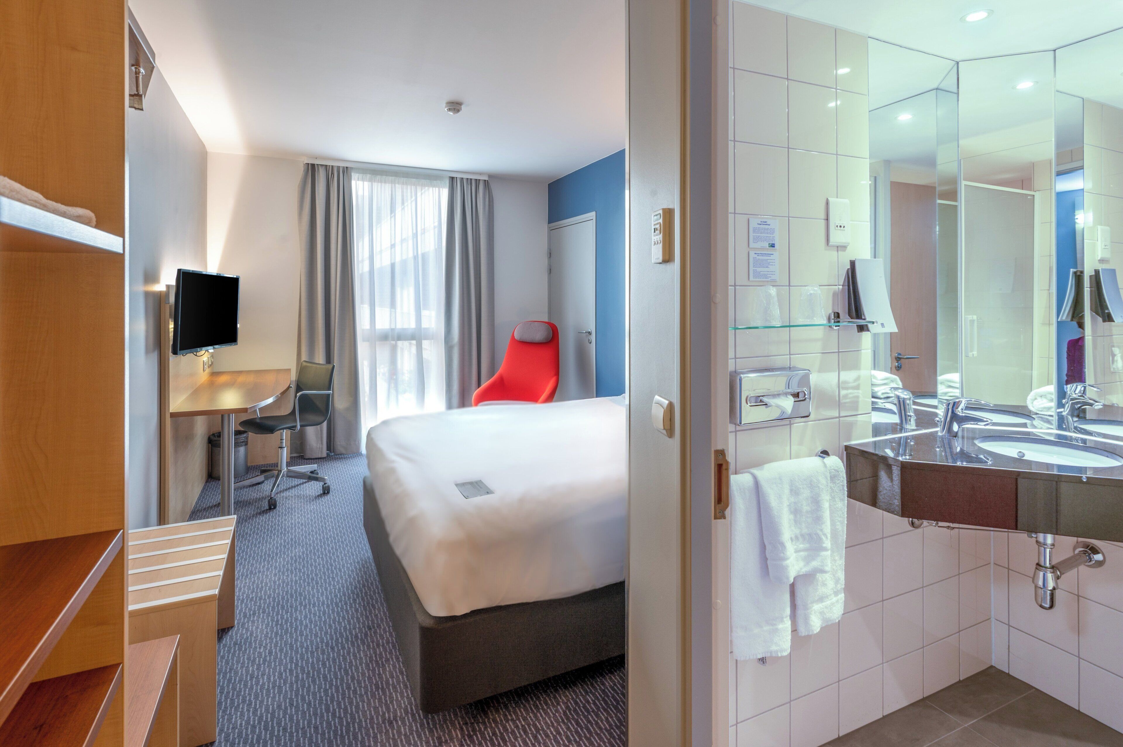 Ein Hotelzimmer mit Bett, Schreibtisch, Stuhl, Fernseher und einem Badezimmer mit Waschbecken und Spiegel.
