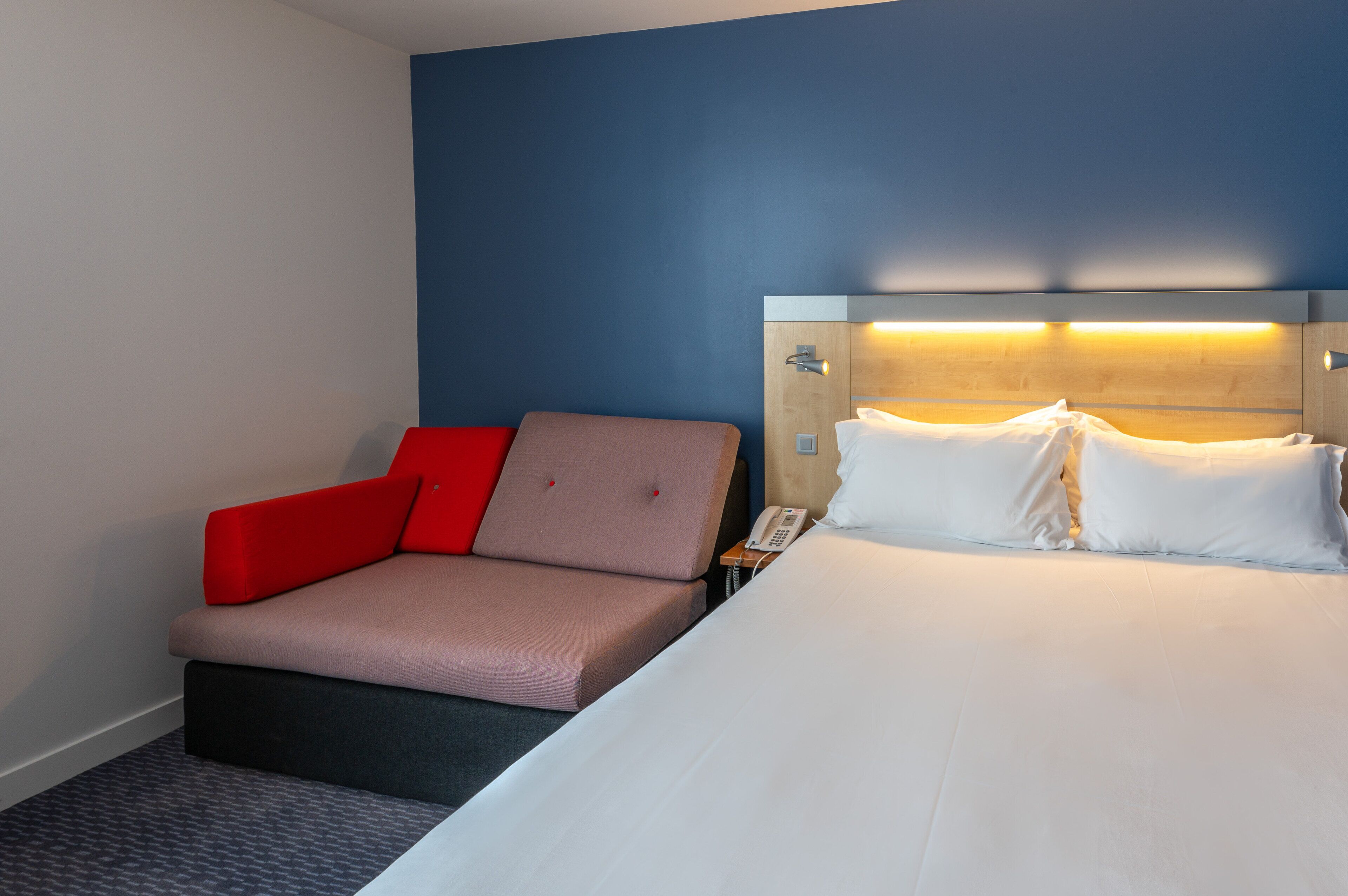 Habitación de hotel con cama, sofá rojo y color beige, mesita de noche con teléfono y pared con detalle azul.