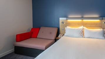 Habitación de hotel con cama, sofá rojo y color beige, mesita de noche con teléfono y pared con detalle azul.